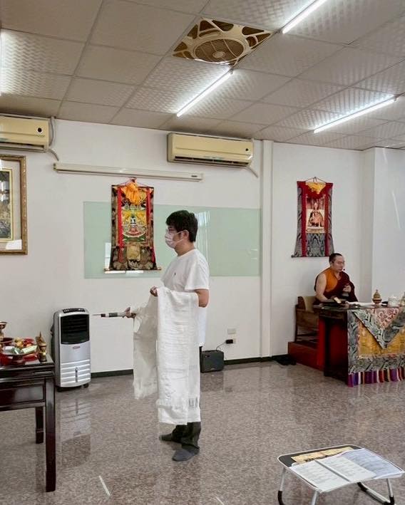 白色室內,一位參與者站在供桌前,另一人坐於旁側。