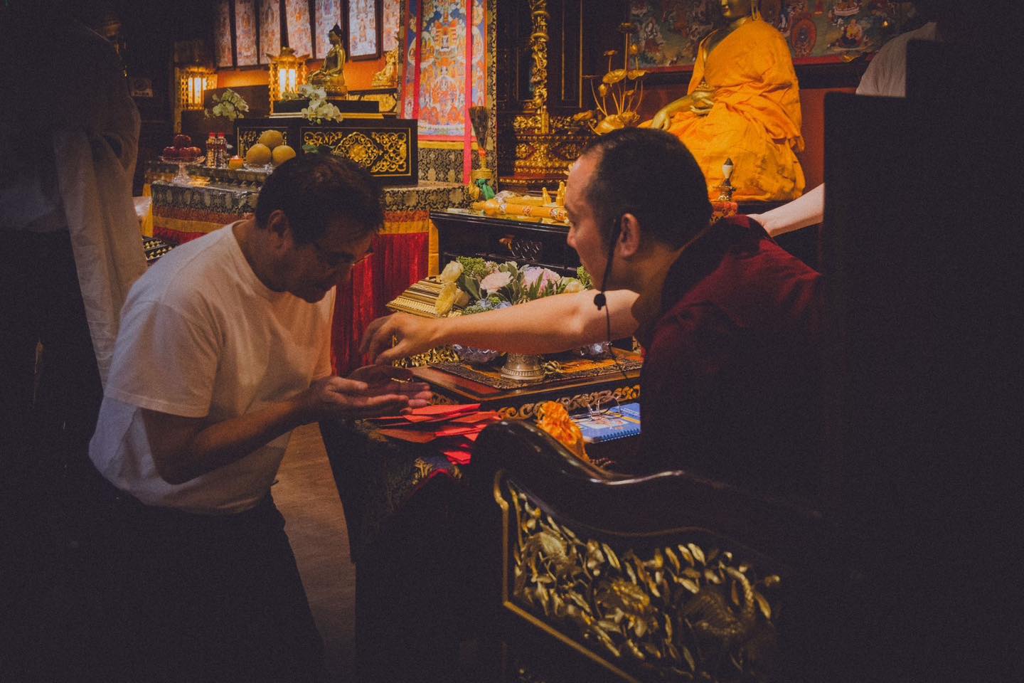 白衣參與者低頭捧手，坐在雕花座椅上的穿袈裟者伸手在其掌心上方放置小物。