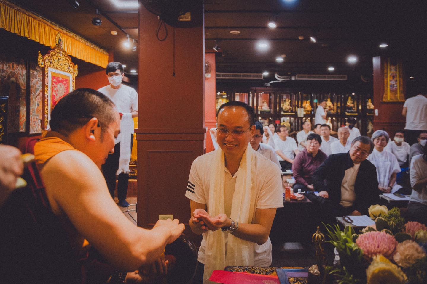 白衣參與者面向穿袈裟者張開雙手，後方多位參與者坐在佛堂內觀看。