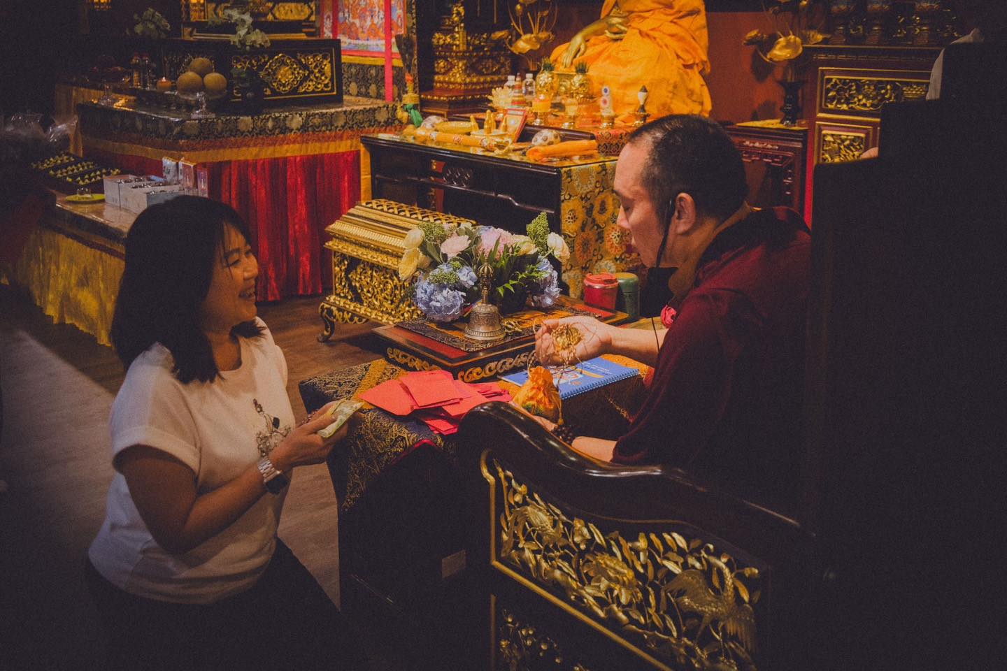 白衣參與者跪坐在雕花座椅前，手持鈔票，穿袈裟者手上拿著一串金色物件。